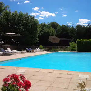 Blauer Camping-Pool mit Liegen, Sonnenschirmen und gepflasterter Terrasse, umgeben von Hecken und Bäumen.