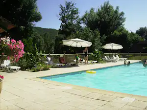 Schwimmbecken auf einem Campingplatz mit Sonnenliegen, Sonnenschirmen und gepflegtem Garten, im Hintergrund Bäume und Hügel.