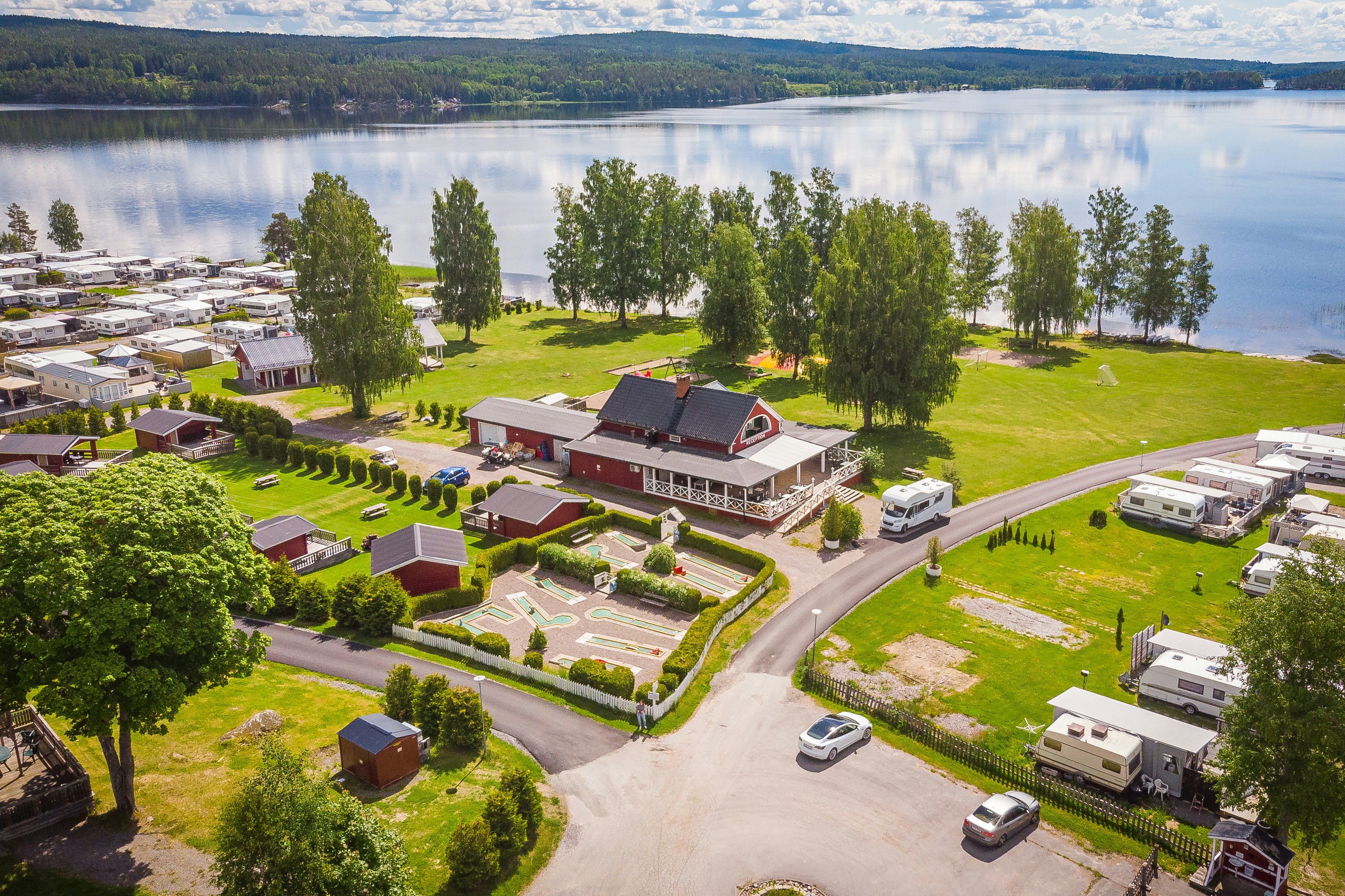 Luftaufnahme eines Campingplatzes mit Wohnwagen, kleinen Häusern und Rasenflächen am See, mit Zufahrtsstraße und Bäumen.