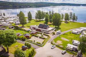Luftaufnahme eines Campingplatzes mit Wohnwagen, kleinen Häusern und Rasenflächen am See, mit Zufahrtsstraße und Bäumen.