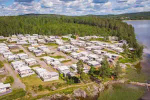 Luftaufnahme eines Campingplatzes mit vielen Stellplätzen, Wohnwagen und Mobilheimen zwischen Wegen, Bäumen und einem kleinen Gewässer.