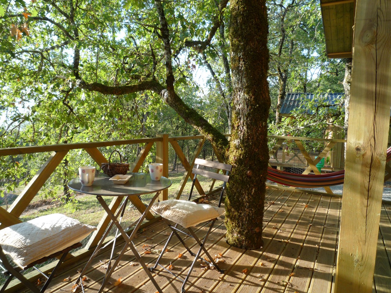 Holzterrasse am Campingplatz mit Tisch und Stühlen, daneben ein Hängesessel und Bäume im Hintergrund.