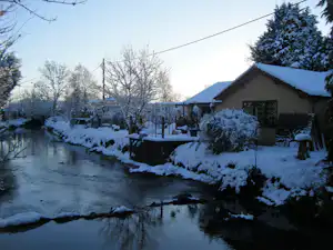 Verschneiter Fluss mit Ufer im Garten, daneben ein Haus mit Satteldach und Hecke, Bäume und Zaun im Hintergrund.