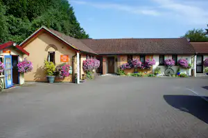 Campinggebäude mit Eingang, Blumenampeln und gepflastertem Hof, umgeben von Bäumen und grünen Hecken.