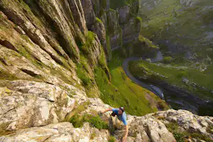 Bergsteiger am Felsvorsprung mit Blick auf einen schmalen Bergpfad und eine kurvige Straße im grünen Tal.
