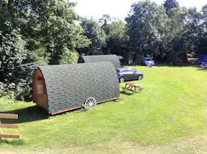 Kleines Campingfahrzeug auf einer grünen Wiese, daneben ein Auto und Gartenmöbel, umgeben von Bäumen.