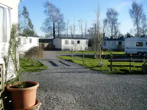 Schotterweg auf dem Campingplatz zwischen Wohnwagen und Gebäuden, mit Hecken, Bäumen und einem Topf am Zeltbereich.