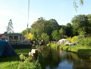 Zelt am Ufer eines ruhigen Flusses, daneben ein Wohnwagen und ein kleines Gebäude, umgeben von Bäumen und Rasen.