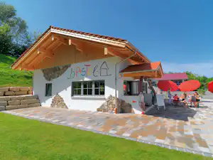 Campinggebäude mit Holzgiebel, großen Fenstern und Terrasse, davor Pflasterweg und rote Sonnenschirme.