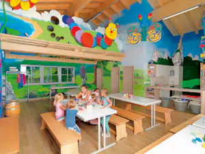 Kinderbereich mit buntem Wandbild, großen Fenstern und Mal-Tischen unter einer Holzdecke.
