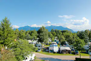 Campingplatz mit Wohnmobilen und Zelten zwischen Bäumen, im Hintergrund Berge und ein blauer Himmel mit Wolken.