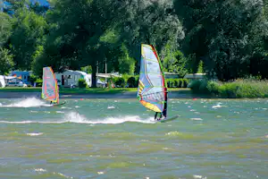 Surfer mit Segel auf dem Wasser, im Hintergrund Wohnwagen, Zelte und Bäume am Campingplatz.