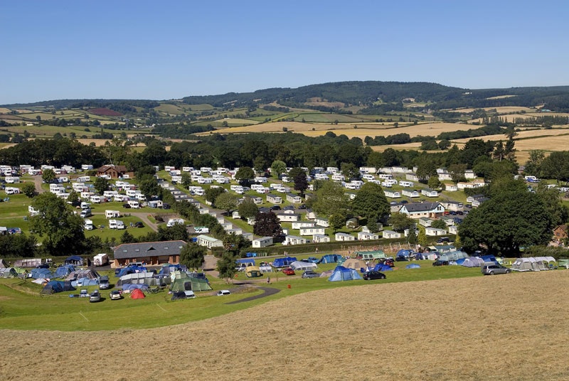 Panoramablick auf einen Campingplatz mit vielen Stellplätzen, Zelten und Wohnwagen, umgeben von Bäumen und grünen Hügeln.