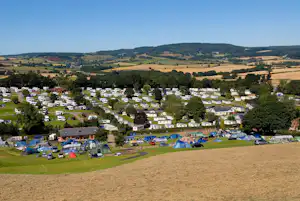 Panoramablick auf einen Campingplatz mit vielen Stellplätzen, Zelten und Wohnwagen, umgeben von Bäumen und grünen Hügeln.