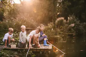 Familie auf einem Steg am Fluss, Kinder mit Kescher und Eimer, im Hintergrund Bäume und Ufervegetation.