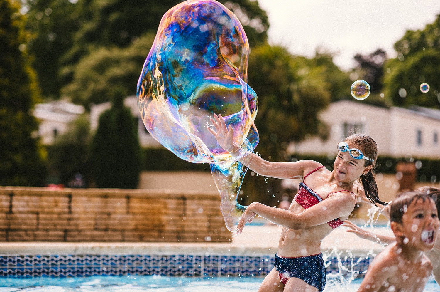Kinder und Erwachsene spielen im Campingpool mit großen Seifenblasen, im Hintergrund Bäume und ein Gebäude.