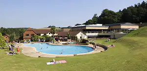 Großer Außenpool auf einem Campingplatz, umgeben von Rasen, Liegen und einem Gebäude mit Terrasse im Hintergrund.