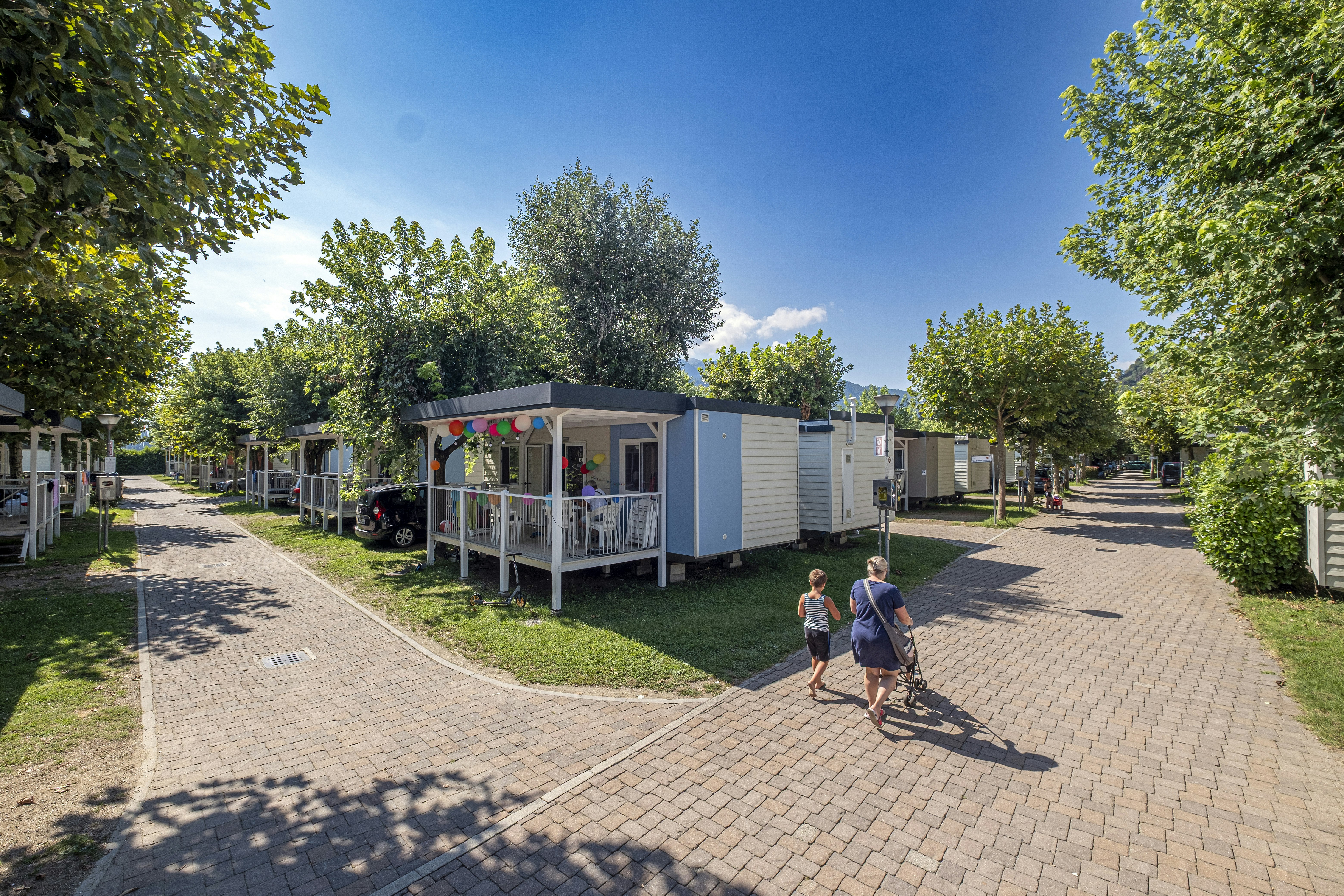 Campingplatz mit Wohnwagen und Stellplätzen auf gepflastertem Weg, Bäume und kleine Gebäude im Hintergrund.