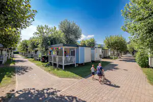 Campingplatz mit Wohnwagen und Stellplätzen auf gepflastertem Weg, Bäume und kleine Gebäude im Hintergrund.