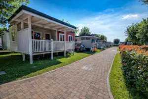 Stellplatz mit Wohnwagen und Holzveranda, gepflasterter Weg, Rasen und Hecken, im Hintergrund weitere Unterkünfte und Bäume.