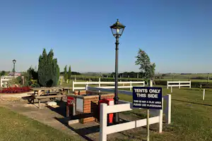 Terrasse mit Holztisch und Bänken, Laternenmast und Zaun; Schild „TENTS ONLY“ vor grünen Feldern und Bäumen.