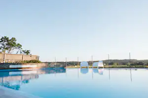 Campingpool mit blauem Beckenrand, zwei weißen Liegestühlen und Zaun im Hintergrund, dahinter Wiese und Bäume.