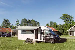 Wohnmobil mit Markise auf dem Campingplatz, davor Tisch und Stühle, im Hintergrund Bungalows und Bäume.
