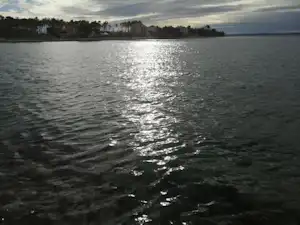 Ruhiger See mit spiegelnden Lichtreflexen, Uferlinie mit Bäumen und Häusern in der Ferne, Blick über das Wasser.
