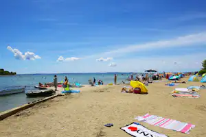 Badeufer am See mit vielen Zelten, Strandliegen und Booten im Wasser, im Hintergrund Menschen und Uferpromenade.