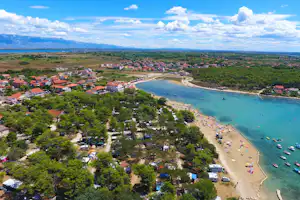 Luftaufnahme eines Campingplatzes an der Küste mit Strand, Booten im Wasser und vielen Stellplätzen zwischen Bäumen.