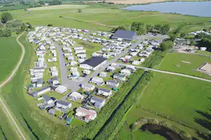 Luftaufnahme eines Campingplatzes mit vielen Wohnwagen und Mobilheimen, Zufahrtsstraße, Hecken und Feldern am See.