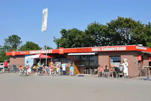 Einkaufsmarkt mit orangefarbenem Dach, Fahrradständern und mehreren Fahrrädern vor dem Gebäude, Bäume im Hintergrund.