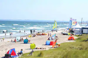 Strandcamping am Meer: Zelte, Strandstühle und Sonnenschirme auf dem Kiesstrand, im Hintergrund Segelboote und viele Badegäste.