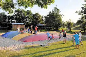 Kinder spielen auf einem bunten Wasserspielplatz mit Rutsche, im Hintergrund Bäume und ein Gebäude am Campingplatz.