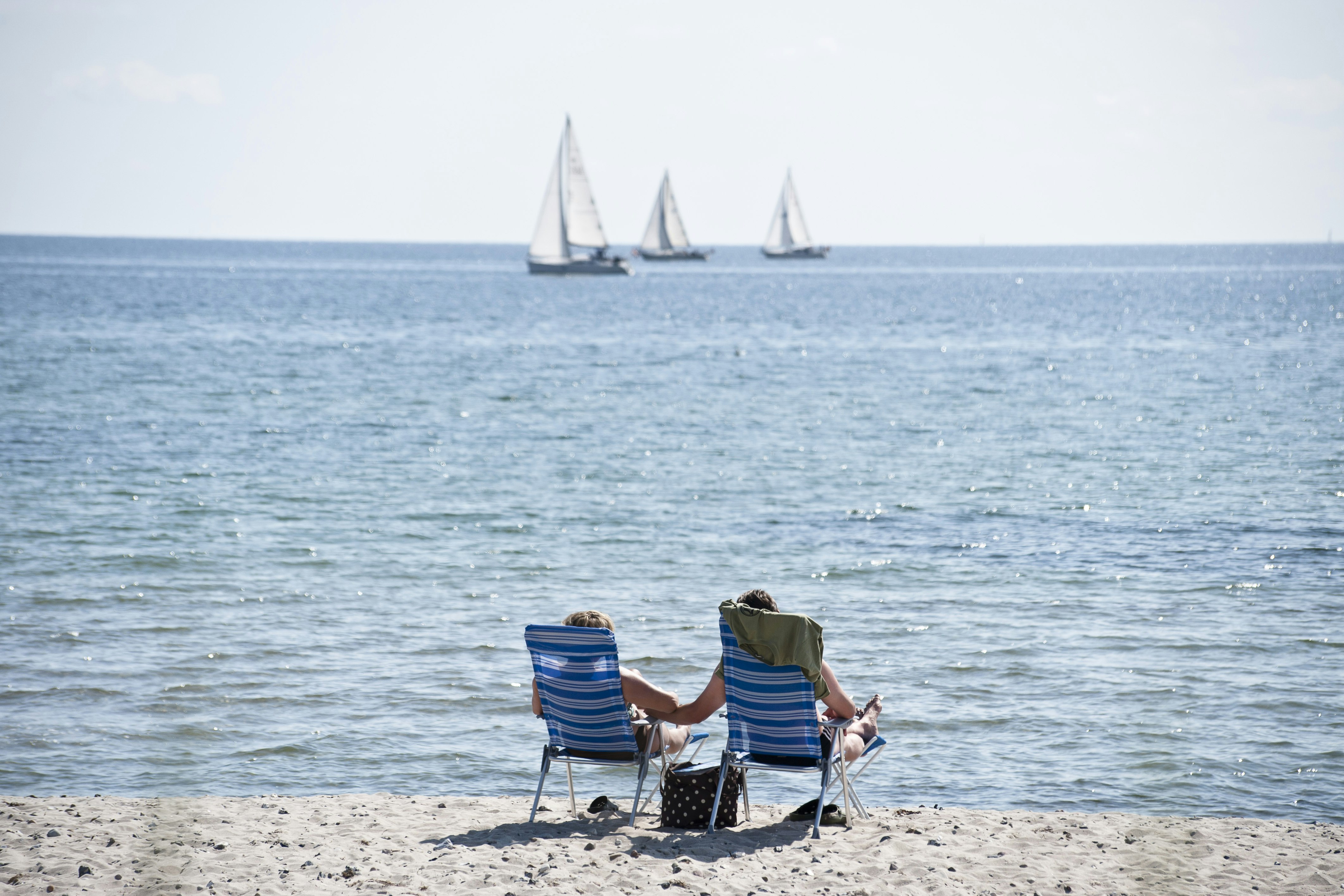 Zwei Campingstühle am Strand vor dem Meer, im Hintergrund Segelboote auf ruhiger Wasserfläche.