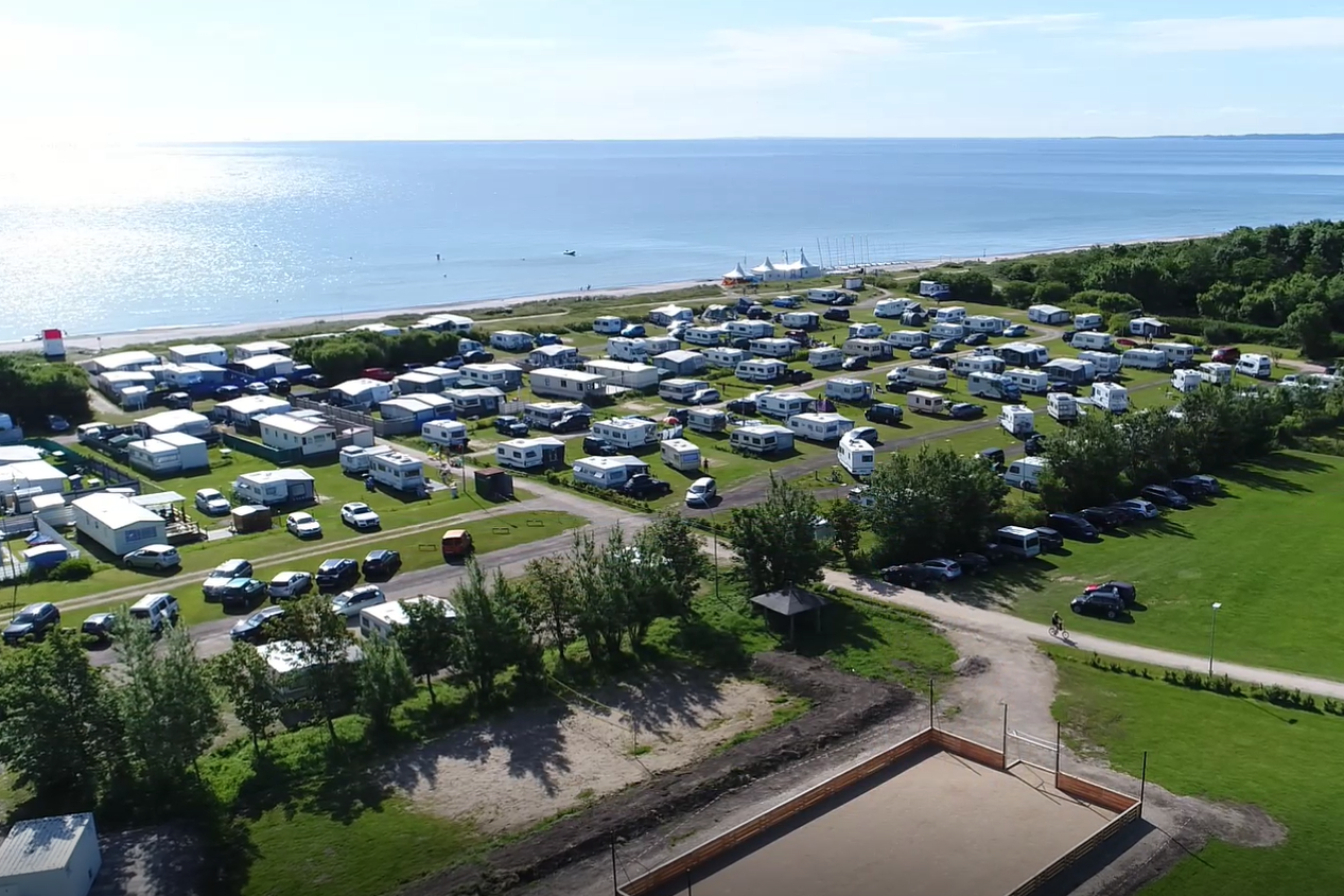 Luftaufnahme eines Campingplatzes mit vielen Stellplätzen, grünen Wiesen und Blick auf das Meer.