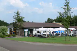 Camping-Eingang mit Steingebäude, grünen Toren und weißen Zelten hinter einem Zaun, umgeben von Bäumen und Rasen.