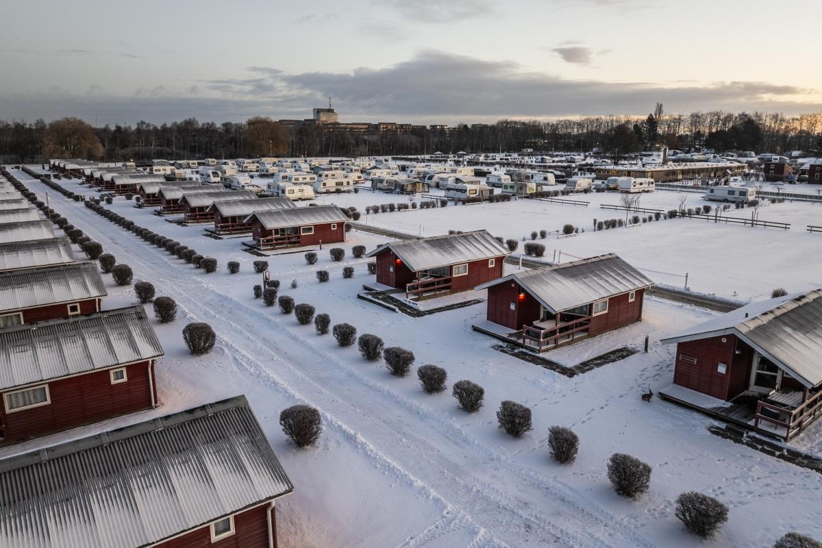 Campingplatz mit roten Holzhütten und überdachten Terrassen im Schnee, Reihen von Unterkünften und verschneiten Wegen.