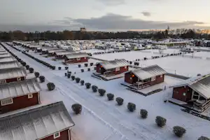 Campingplatz mit roten Holzhütten und überdachten Terrassen im Schnee, Reihen von Unterkünften und verschneiten Wegen.