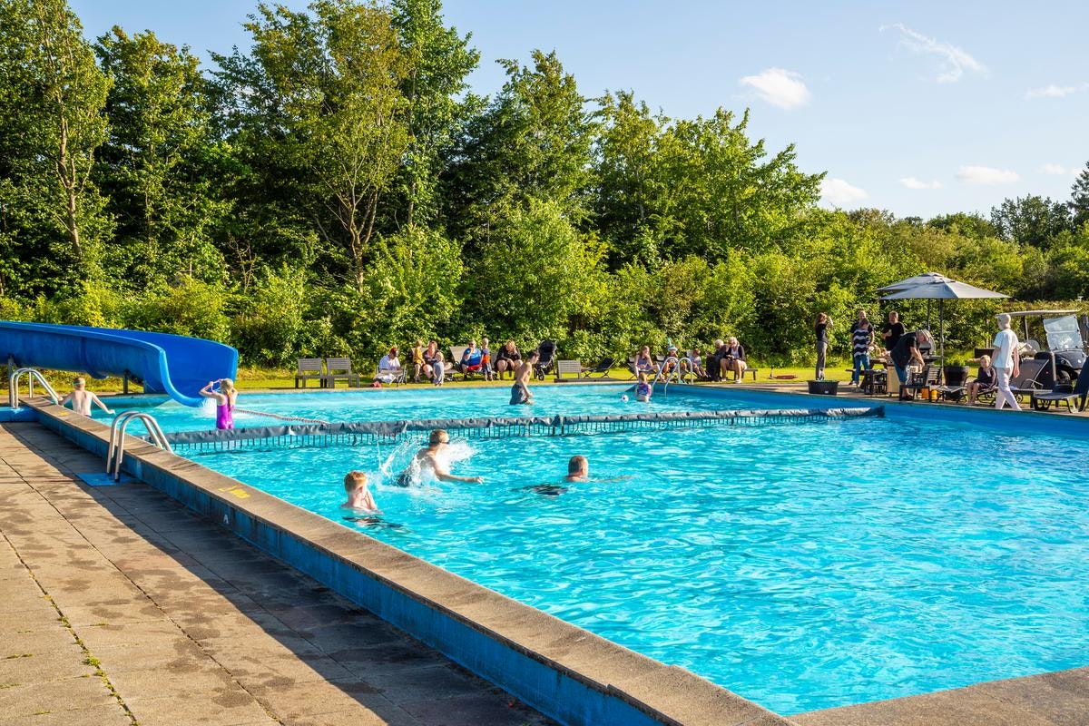 Großes Camping-Schwimmbecken mit blauer Wasserfläche, Liegen und mehreren Personen am Beckenrand vor Bäumen und Sonnenschirmen.