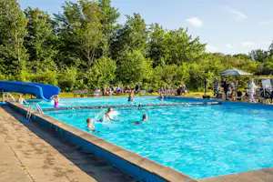 Großes Camping-Schwimmbecken mit blauer Wasserfläche, Liegen und mehreren Personen am Beckenrand vor Bäumen und Sonnenschirmen.