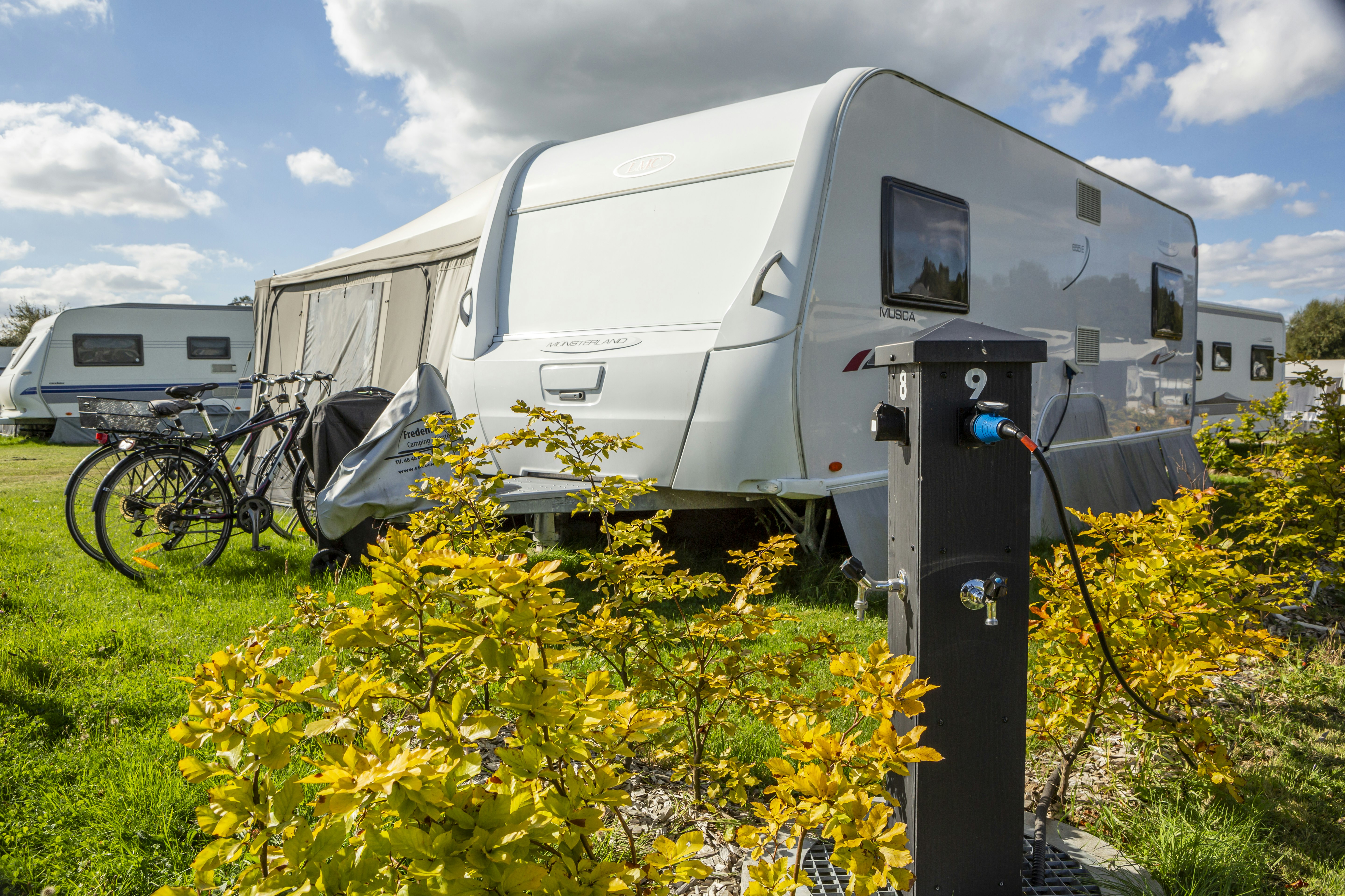 Wohnwagen auf dem Campingplatz mit Stromanschluss und Kabel, im Vordergrund gelbe Sträucher und grünes Gras.