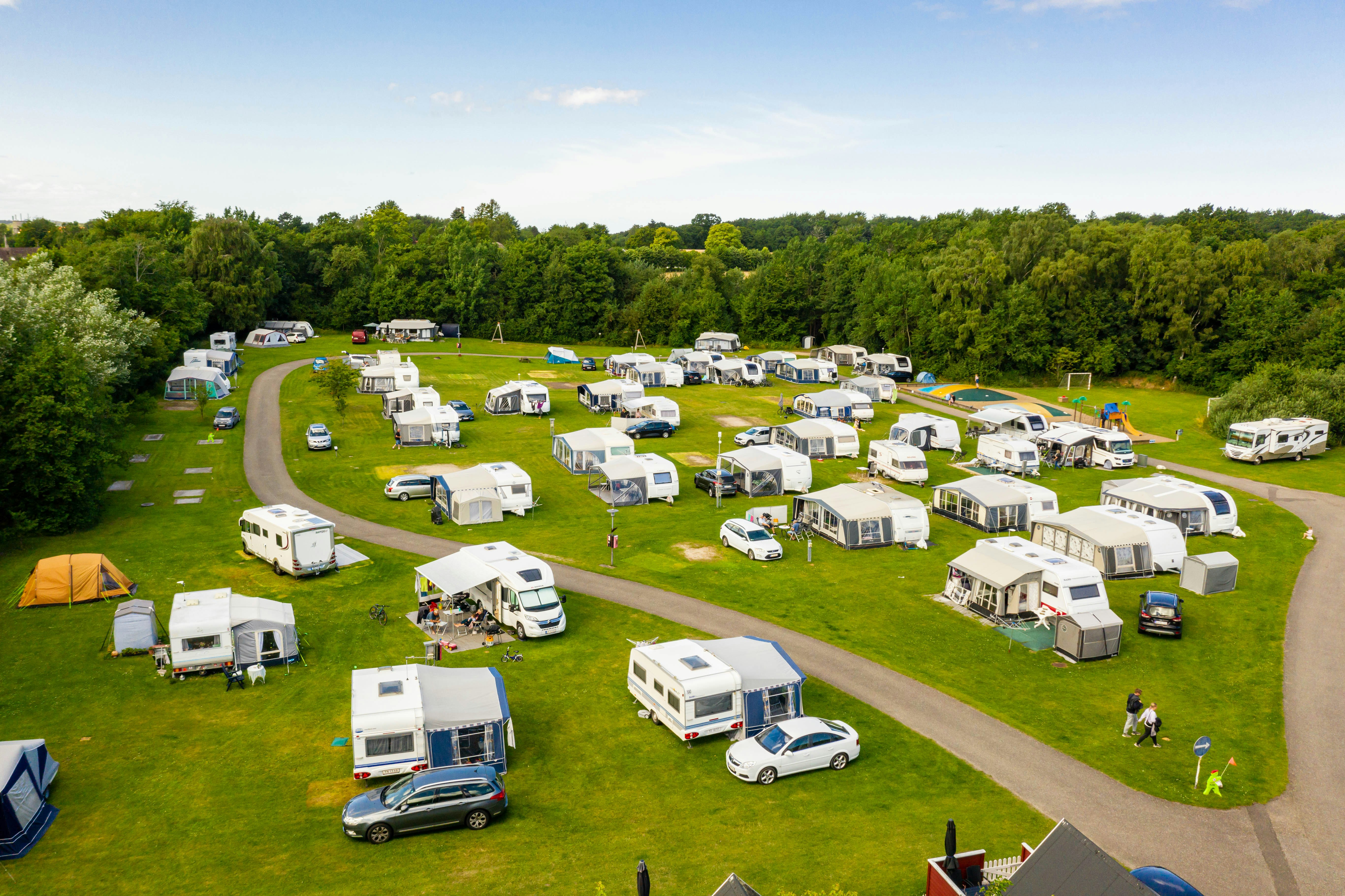 Luftaufnahme eines Campingplatzes mit Wohnwagen und Zelten auf grünen Stellplätzen, Wegen und Bäumen im Hintergrund.