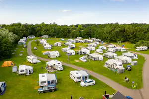Luftaufnahme eines Campingplatzes mit Wohnwagen und Zelten auf grünen Stellplätzen, Wegen und Bäumen im Hintergrund.