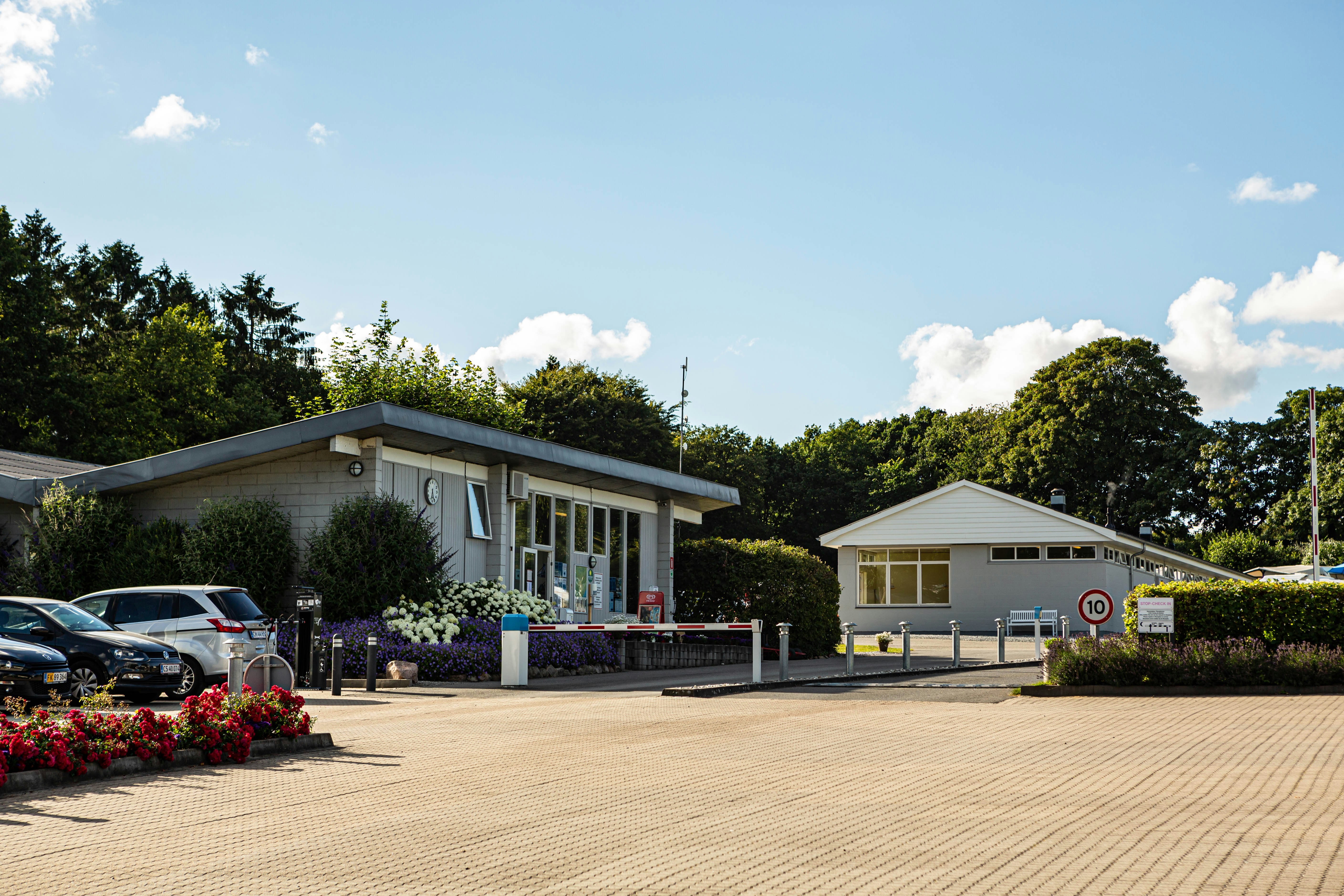 Eingangsgebäude eines Campingplatzes mit Parkplatz, Blumenbeeten und Hecken vor dem Gebäude unter blauem Himmel.