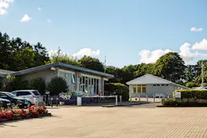 Eingangsgebäude eines Campingplatzes mit Parkplatz, Blumenbeeten und Hecken vor dem Gebäude unter blauem Himmel.