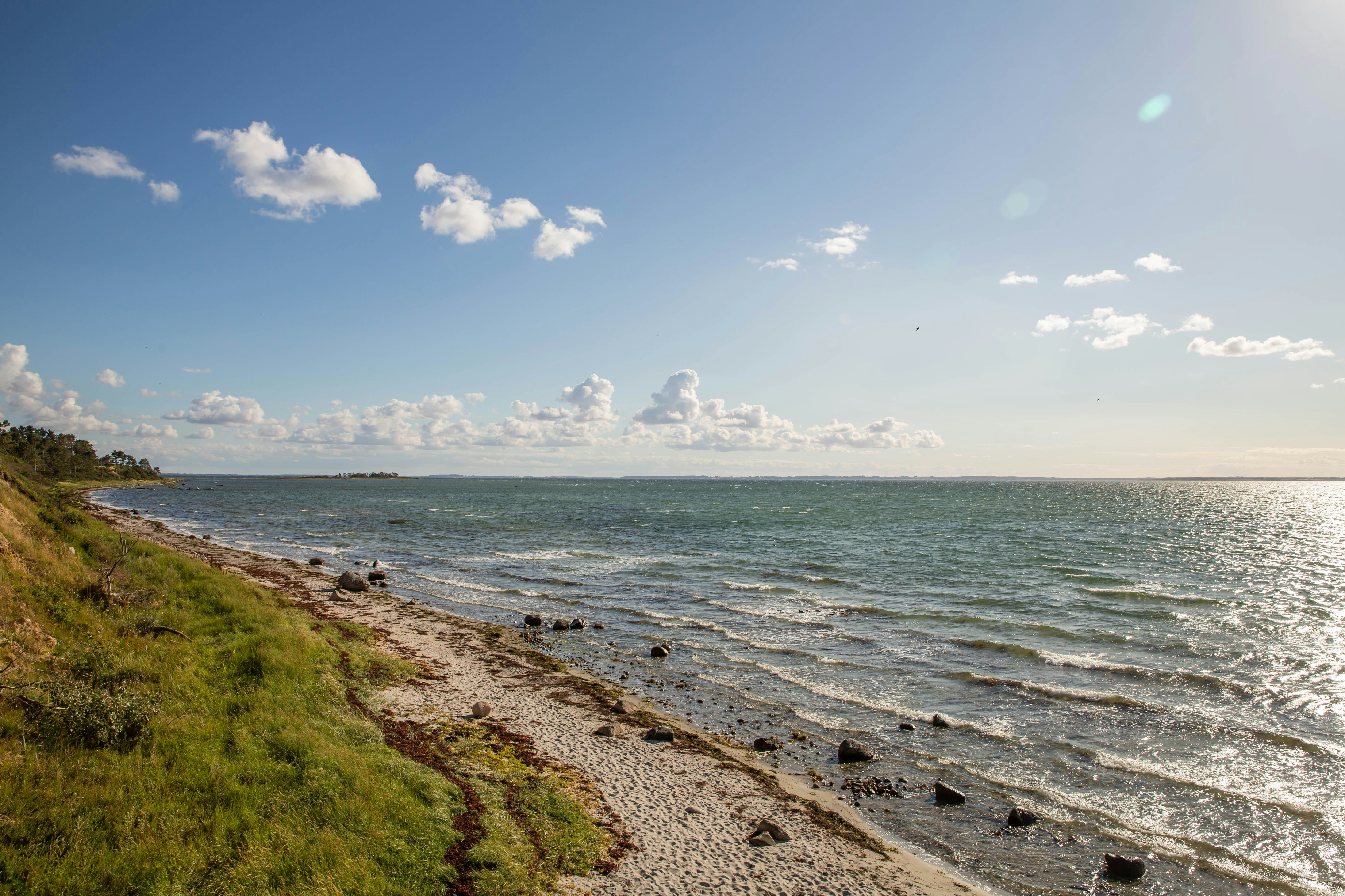 Küstenlinie mit Kiesstrand, Wellen und grünem Uferweg, Blick auf das Meer und den Himmel mit Wolken.