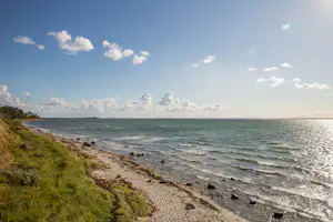 Küstenlinie mit Kiesstrand, Wellen und grünem Uferweg, Blick auf das Meer und den Himmel mit Wolken.