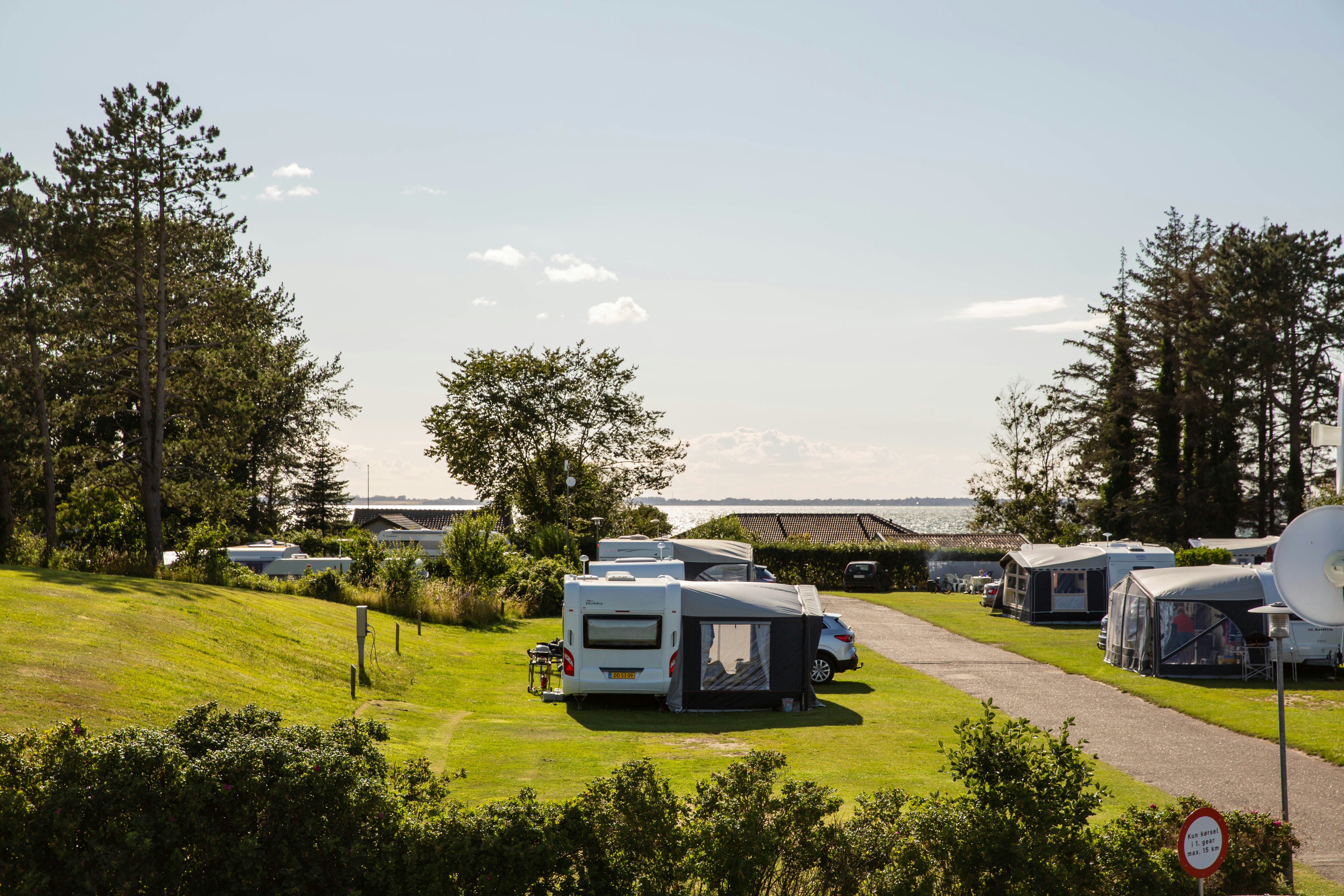 Stellplätze mit Wohnwagen und Vorzelten auf einer grünen Wiese, Hecken und Bäume am Rand, Weg zur Rezeption.