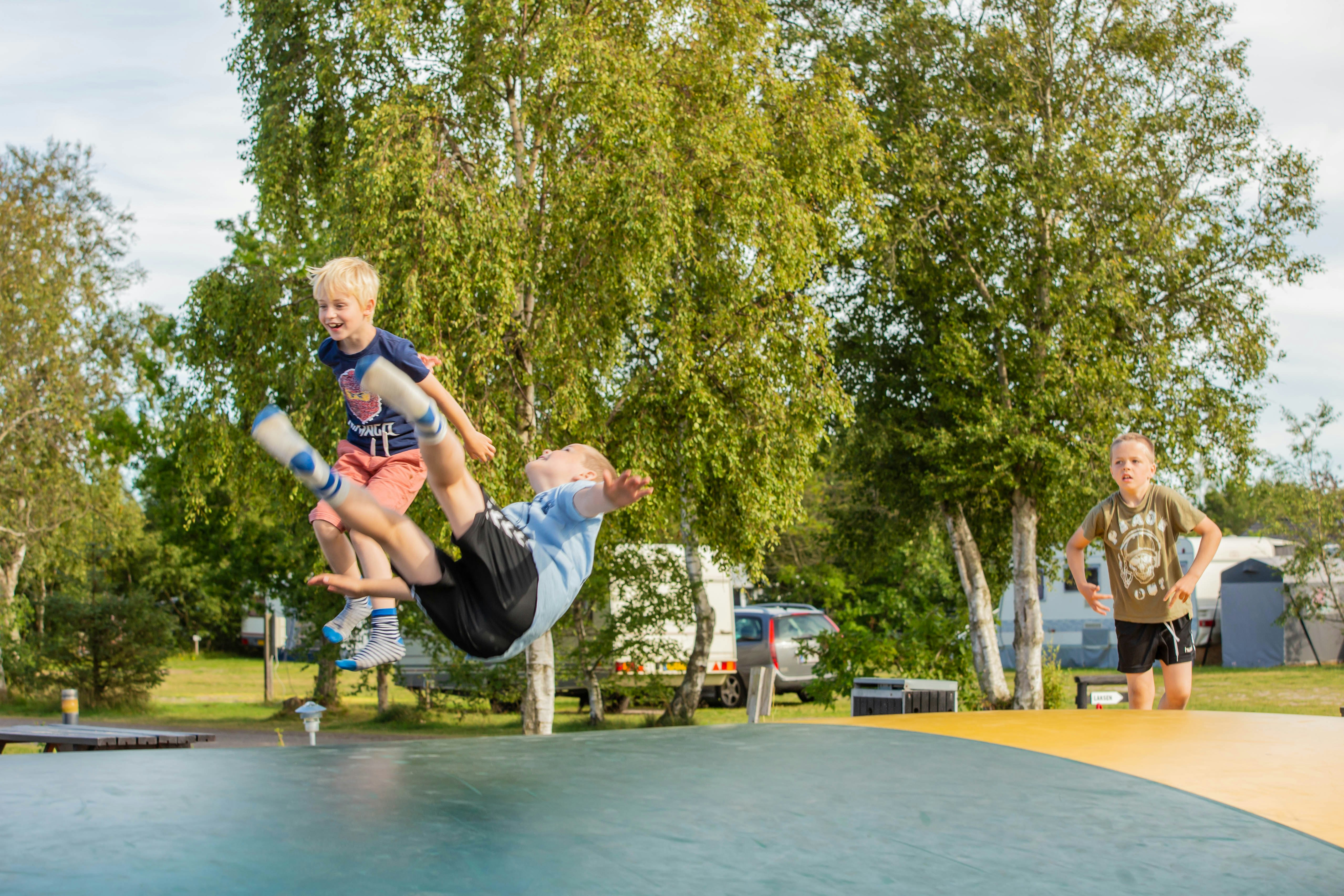 Kinder springen auf einem Trampolin im Campinggelände, im Hintergrund Bäume, Wohnwagen und ein Tisch.